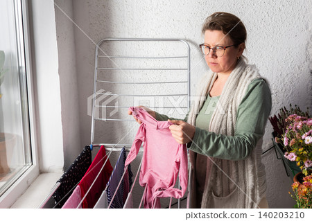 A 50-year-old woman is hanging her laundry on a metal drying rack on her balcony. She looks focused while handling the clothes and has flowers nearby 140203210