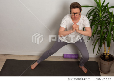 A woman aged 52 performs a yoga stretch in her home. She stands on a mat and focuses on her form. The room has a plain wall and a green plant nearby A woman aged 52 performs a yoga stretch in her home. She stands on a mat and focuses on her form. The room has a plain wall and a green plant nearby 140203356
