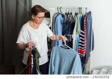 Woman sorts and hangs washed clothes on a hanger. She holds a blue sweatshirt and several other items in her hands. This scene takes place in a home environment Woman sorts and hangs washed clothes on a hanger. She holds a blue sweatshirt and several other items in her hands. This scene takes place in a home environment 140203358