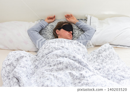 A 50-year-old woman is comfortably sleeping in her bed with a sleep mask on. She is wrapped in a soft blanket, resting peacefully in a serene bedroom setting A 50-year-old woman is comfortably sleeping in her bed with a sleep mask on. She is wrapped in a soft blanket, resting peacefully in a serene bedroom setting 140203520