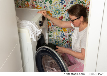 A woman takes clean clothes out of a washing machine in a laundry room. She is focused on her task, surrounded by a colorful wall. It is daytime and chores are being done A woman takes clean clothes out of a washing machine in a laundry room. She is focused on her task, surrounded by a colorful wall. It is daytime and chores are being done 140203523