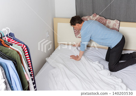 A 53-year-old man is changing the bed linen in his bedroom. He is pulling the fresh linen over the mattress while organizing the space for the day A 53-year-old man is changing the bed linen in his bedroom. He is pulling the fresh linen over the mattress while organizing the space for the day 140203530