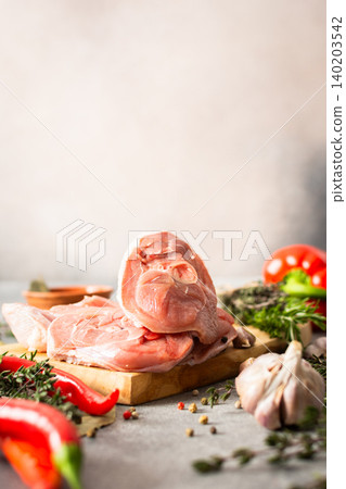 Turkey meat is placed on a wooden cutting board in a kitchen. Fresh herbs, garlic, and colorful vegetables surround the meat, adding to the cooking scene 140203542