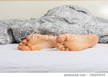 A man enjoys his time relaxing on a bed. His feet can be seen sticking out from under a gray blanket. It looks like a comfortable setting at home or in a hotel A man enjoys his time relaxing on a bed. His feet can be seen sticking out from under a gray blanket. It looks like a comfortable setting at home or in a hotel 140203545