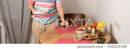 A 53-year-old woman sets the table for dinner. She carries a stack of plates and a glass of water. Fresh fruits and vegetables are arranged on the table, ready for the meal, banner A 53-year-old woman sets the table for dinner. She carries a stack of plates and a glass of water. Fresh fruits and vegetables are arranged on the table, ready for the meal, banner 140203580