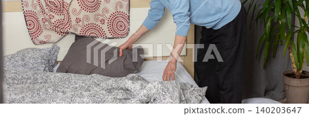 A 53-year-old man replaces the bed linen in his bedroom. He gathers clean sheets and works on making the bed comfortable for sleeping, banner A 53-year-old man replaces the bed linen in his bedroom. He gathers clean sheets and works on making the bed comfortable for sleeping, banner 140203647