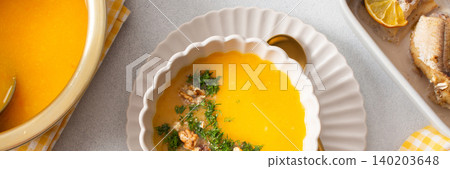 Pumpkin cream soup sits in a bowl with toppings next to a dish of baked hake and lemon. A spoon rests beside the soup in a clean dining area, banner Pumpkin cream soup sits in a bowl with toppings next to a dish of baked hake and lemon. A spoon rests beside the soup in a clean dining area, banner 140203648