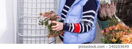 A 50-year-old woman tends to autumn flowers on her balcony. She focuses on each plant with care while surrounded by city buildings. It is a cool afternoon in the fall season, banner 140203649