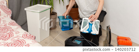 A 53-year-old man is getting ready to clean by preparing a vacuum cleaner. He focuses on the vacuum's parts in a plain room with plants nearby, banner 140203712