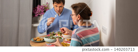 A married couple is having dinner together at home. They sit at a small table near a window with a bowl of salad and other food. The light is warm and cozy in the evening, banner 140203713