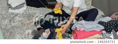 A 52-year-old woman sits on her bed with laundry scattered around her. She focuses on sorting socks and organizing other clothes, performing a daily task at home, banner A 52-year-old woman sits on her bed with laundry scattered around her. She focuses on sorting socks and organizing other clothes, performing a daily task at home, banner 140203731