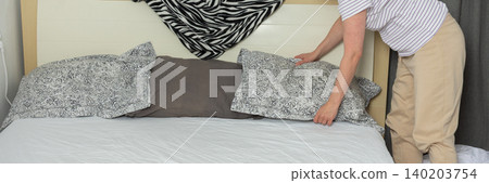 A 53-year-old woman is in a light room. She replaces her old bed linen with new ones. She sits on the bed and adjusts the covers to make it tidy and neat, banner A 53-year-old woman is in a light room. She replaces her old bed linen with new ones. She sits on the bed and adjusts the covers to make it tidy and neat, banner 140203754