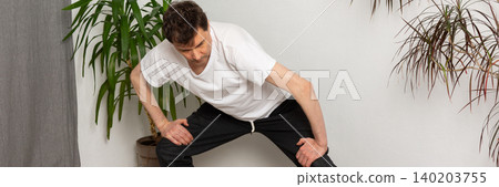 A 53-year-old man performs exercises in his living room at home, stretching and working on his body movements. Green plants are visible in the background adding to the atmosphere, banner A 53-year-old man performs exercises in his living room at home, stretching and working on his body movements. Green plants are visible in the background adding to the atmosphere, banner 140203755