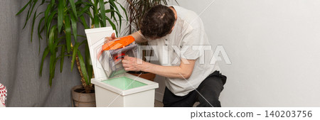 A 53-year-old man is getting ready to clean by preparing a vacuum cleaner. He focuses on the vacuum's parts in a plain room with plants nearby, banner A 53-year-old man is getting ready to clean by preparing a vacuum cleaner. He focuses on the vacuum's parts in a plain room with plants nearby, banner 140203756