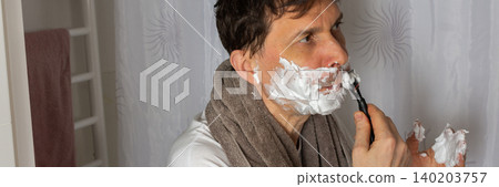 A 53 year old man shaves in the bathroom during his morning routine. He uses a razor and shaving cream while wearing a towel around his neck. Water is visible in the sink, banner A 53 year old man shaves in the bathroom during his morning routine. He uses a razor and shaving cream while wearing a towel around his neck. Water is visible in the sink, banner 140203757