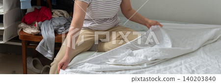 A 53-year-old woman is working in her bedroom. She is changing the bed linen and placing new sheets on the bed. Clothes are seen nearby in the room, banner A 53-year-old woman is working in her bedroom. She is changing the bed linen and placing new sheets on the bed. Clothes are seen nearby in the room, banner 140203840