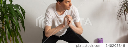 A 53-year-old man practices exercises at home on a yoga mat. He focuses on his strength and flexibility while avoiding distractions from the surroundings. Plants stand around him, banner A 53-year-old man practices exercises at home on a yoga mat. He focuses on his strength and flexibility while avoiding distractions from the surroundings. Plants stand around him, banner 140203846