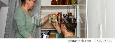 Married couple places jars of canned goods onto shelves in their pantry. They are inside their home with a view of flowers and the city from the balcony. It is autumn, banner Married couple places jars of canned goods onto shelves in their pantry. They are inside their home with a view of flowers and the city from the balcony. It is autumn, banner 140203849