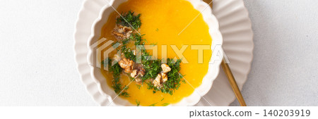 A bowl of pumpkin cream soup sits on a plate. The soup contains nuts and herbs. A yellow checkered napkin lies nearby. This meal can be enjoyed at breakfast or lunch, banner 140203919