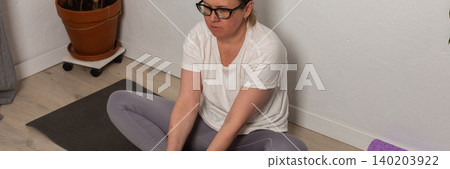 A 52-year-old woman exercises at home, sitting on a yoga mat. She is stretching and holding her feet while focusing on her breathing and posture in a simple indoor setting, banner A 52-year-old woman exercises at home, sitting on a yoga mat. She is stretching and holding her feet while focusing on her breathing and posture in a simple indoor setting, banner 140203922