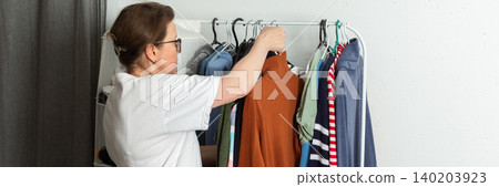 A 52-year-old woman sorts and hangs washed clothes on a hanger inside her home. She is organizing different garments after completing her laundry tasks, banner A 52-year-old woman sorts and hangs washed clothes on a hanger inside her home. She is organizing different garments after completing her laundry tasks, banner 140203923