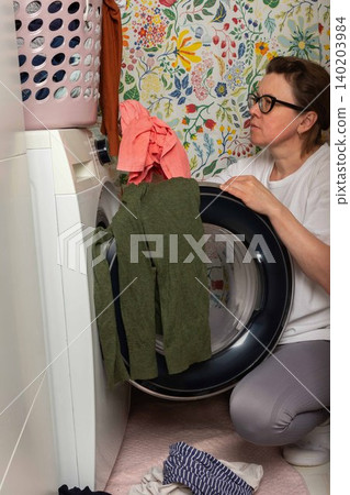 Woman stands in bathroom putting clothes into washing machine while surrounded by towels and laundry, carrying out daily household task with focus and care Woman stands in bathroom putting clothes into washing machine while surrounded by towels and laundry, carrying out daily household task with focus and care 140203984