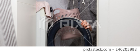 A man is taking clothes out of a washing machine. He is putting them into a basket. This is happening in a laundry room at home. The lighting shows it is daytime, banner 140203998