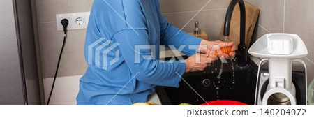 In a bright kitchen, a woman washes carrots at the sink. She focuses on cleaning the vegetables before starting the process of fermenting cabbage, banner In a bright kitchen, a woman washes carrots at the sink. She focuses on cleaning the vegetables before starting the process of fermenting cabbage, banner 140204072