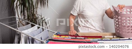 A 50-year-old woman hangs freshly washed laundry on a drying rack inside her room. She is holding a laundry basket filled with clothes, organizing her space, banner A 50-year-old woman hangs freshly washed laundry on a drying rack inside her room. She is holding a laundry basket filled with clothes, organizing her space, banner 140204076