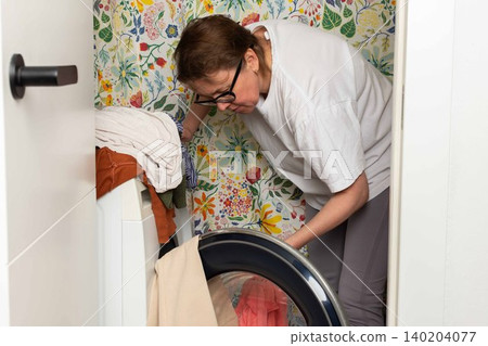 Woman stands in bathroom putting clothes into washing machine while surrounded by towels and laundry, carrying out daily household task with focus and care Woman stands in bathroom putting clothes into washing machine while surrounded by towels and laundry, carrying out daily household task with focus and care 140204077