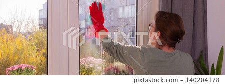 A person cleans windows in a city apartment on a balcony. The scene shows autumn flowers and a clear view outside on a sunny day as winter approaches, banner A person cleans windows in a city apartment on a balcony. The scene shows autumn flowers and a clear view outside on a sunny day as winter approaches, banner 140204178