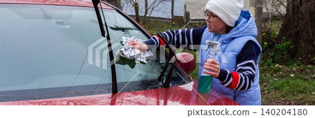 A person is cleaning the windshield of a red car with a cloth while holding a spray bottle. This activity is taking place outside a house on a cloudy day, banner A person is cleaning the windshield of a red car with a cloth while holding a spray bottle. This activity is taking place outside a house on a cloudy day, banner 140204180