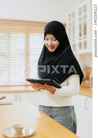 Young Arab Muslim woman in hijab works with tablet computer at home kitchen happy smiling. Young Arab Muslim woman in hijab works with tablet computer at home kitchen happy smiling. 140204223