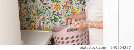 A woman stands in a laundry room holding a basket with clean clothes. She looks focused as she prepares to take out laundry from the washing machine. The room has bright floral wallpaper, banner A woman stands in a laundry room holding a basket with clean clothes. She looks focused as she prepares to take out laundry from the washing machine. The room has bright floral wallpaper, banner 140204257