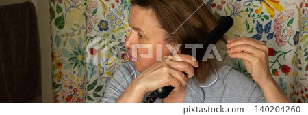 A 50-year-old woman styles her hair with a straightener in a bathroom. Floral curtains add color to the space. She is focused on her hair, preparing for the day ahead, banner A 50-year-old woman styles her hair with a straightener in a bathroom. Floral curtains add color to the space. She is focused on her hair, preparing for the day ahead, banner 140204260
