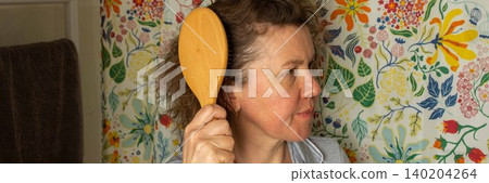 A 50-year-old woman stands in her bathroom holding a wooden hairbrush. She focuses on brushing her hair while facing a floral patterned wall, preparing for her day ahead, banner A 50-year-old woman stands in her bathroom holding a wooden hairbrush. She focuses on brushing her hair while facing a floral patterned wall, preparing for her day ahead, banner 140204264