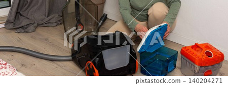 A person is getting ready to vacuum a room. They are attaching a container to the vacuum cleaner while kneeling on the floor. A plant is visible in the background, banner A person is getting ready to vacuum a room. They are attaching a container to the vacuum cleaner while kneeling on the floor. A plant is visible in the background, banner 140204271