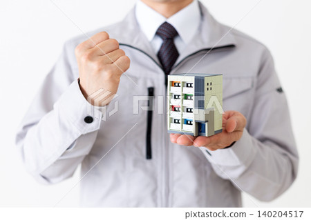 A middle-aged businessman in gray work clothes strikes a triumphant pose while holding a model of an apartment building against a white background. 140204517