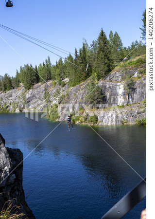 Ruskeala, Russia - July 10, 2021: A man hangs on a bungee above the lake of the Marble Canyon in Ruskeale in Karelia in Russia Ruskeala, Russia - July 10, 2021: A man hangs on a bungee above the lake of the Marble Canyon in Ruskeale in Karelia in Russia 140206274