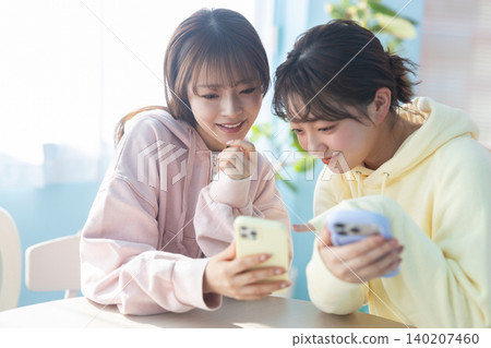 兩位年輕女子在家中互相展示智慧型手機,玩得不亦樂乎。 兩位年輕女子在家中互相展示智慧型手機,玩得不亦樂乎。 140207460