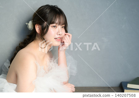 一位身穿婚紗的日本女子麵帶微笑，手托著下巴，背景是灰色的。 140207508