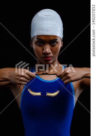 A focused female swimmer holds her swimming goggles in front of her chest, wearing a blue swimsuit and white swim cap, ready for intense competition. A focused female swimmer holds her swimming goggles in front of her chest, wearing a blue swimsuit and white swim cap, ready for intense competition. 140207852