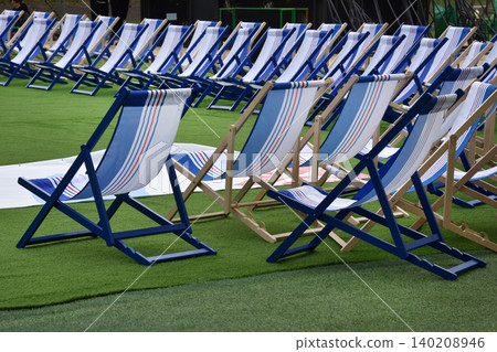 Empty blue and white deck chairs arranged in rows on green turf at an outdoor event. Sun-ready seating evokes summer vacation and holidays like Independence Day and Labor Day. Empty blue and white deck chairs arranged in rows on green turf at an outdoor event. Sun-ready seating evokes summer vacation and holidays like Independence Day and Labor Day. 140208946