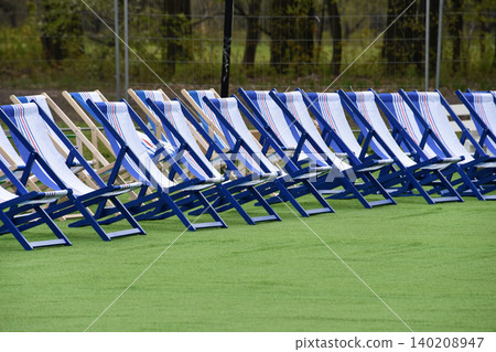 Empty blue and white deck chairs arranged in rows on green turf at an outdoor event. Sun-ready seating evokes summer vacation and holidays like Independence Day and Labor Day. Empty blue and white deck chairs arranged in rows on green turf at an outdoor event. Sun-ready seating evokes summer vacation and holidays like Independence Day and Labor Day. 140208947