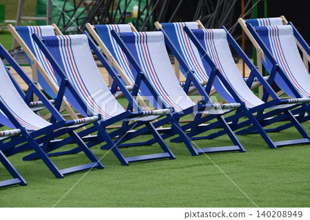 Empty blue and white deck chairs arranged in rows on green turf at an outdoor event. Sun-ready seating evokes summer vacation and holidays like Independence Day and Labor Day. 140208949