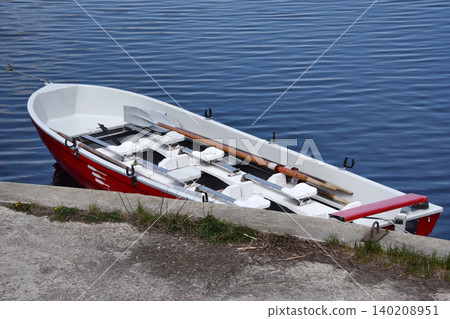 Red rowboat with wooden oars floats on calm blue water at a lakeside. Leisure craft with seats and oars suggests summer holidays and peaceful relaxation. Red rowboat with wooden oars floats on calm blue water at a lakeside. Leisure craft with seats and oars suggests summer holidays and peaceful relaxation. 140208951