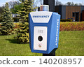 Emergency call box with camera and flashing light mounted on a pole beside a grassy park and campus building. A blue alarm station provides safety and emergency communication 140208957