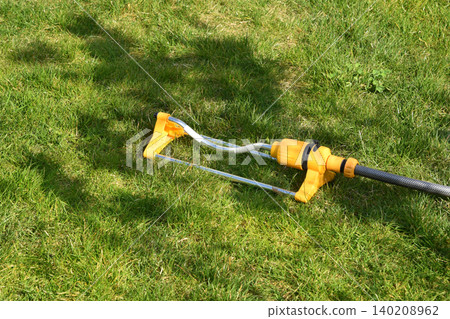 A yellow lawn sprinkler waters grass in a backyard. The sprinkler irrigates the green lawn, capturing spring garden care. A yellow lawn sprinkler waters grass in a backyard. The sprinkler irrigates the green lawn, capturing spring garden care. 140208962
