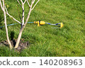 A yellow lawn sprinkler waters grass around a small tree in a backyard. The sprinkler irrigates the green lawn near the tree, capturing spring garden care. 140208963