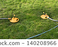 Two yellow oscillating sprinklers water a lush green lawn in a backyard garden. Backyard garden sprinklers irrigate grass on a bright summer day, evoking spring and summer holidays. 140208966